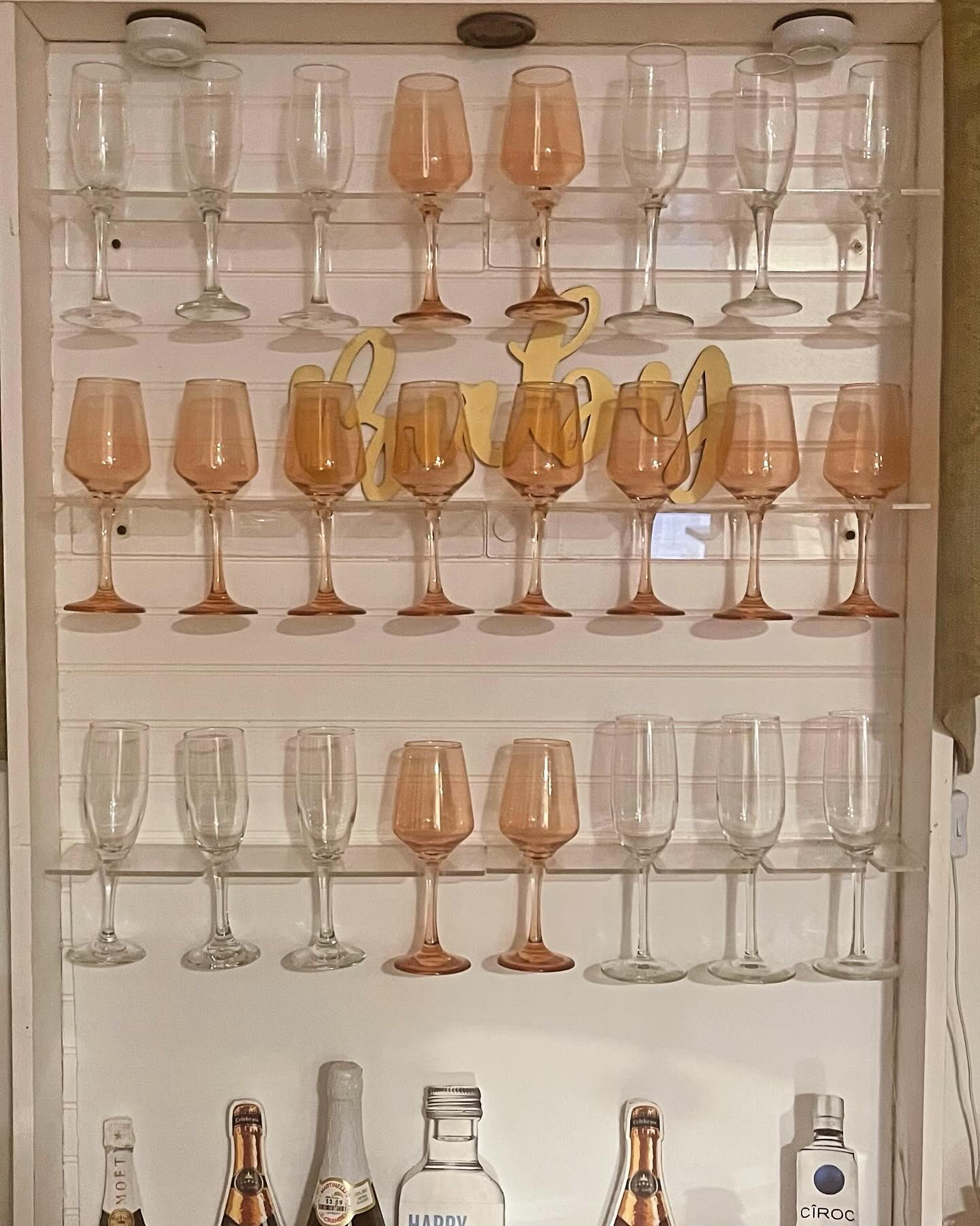 A wine and champagne glass display shelf holding rows of clear and pink-tinted stemware, a gold script sign, and mini liquor bottles