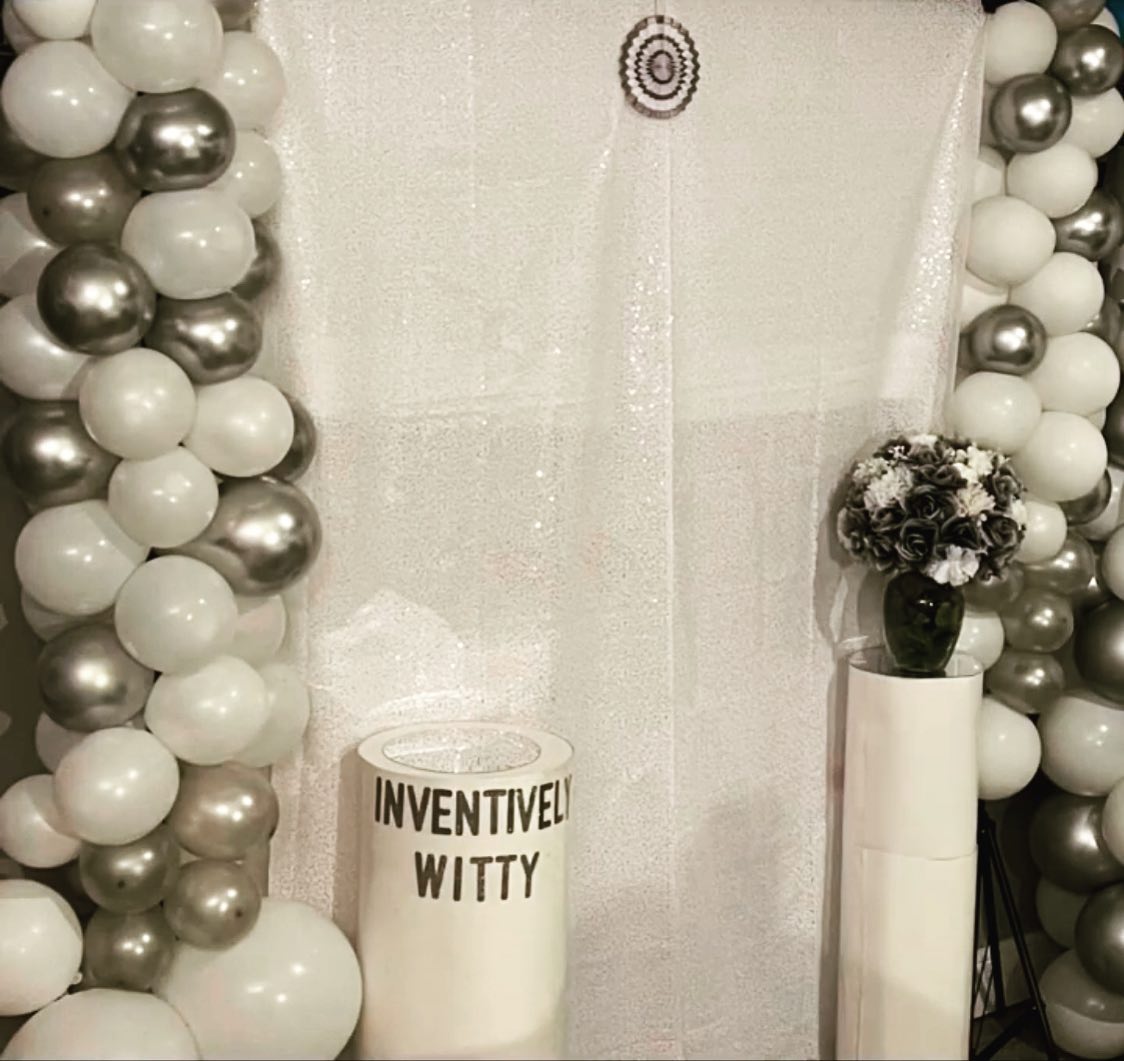 White fabric backdrop flanked by white and silver balloon garlands, with a pedestal reading Inventively Witty and another holding a dark floral arrangement
