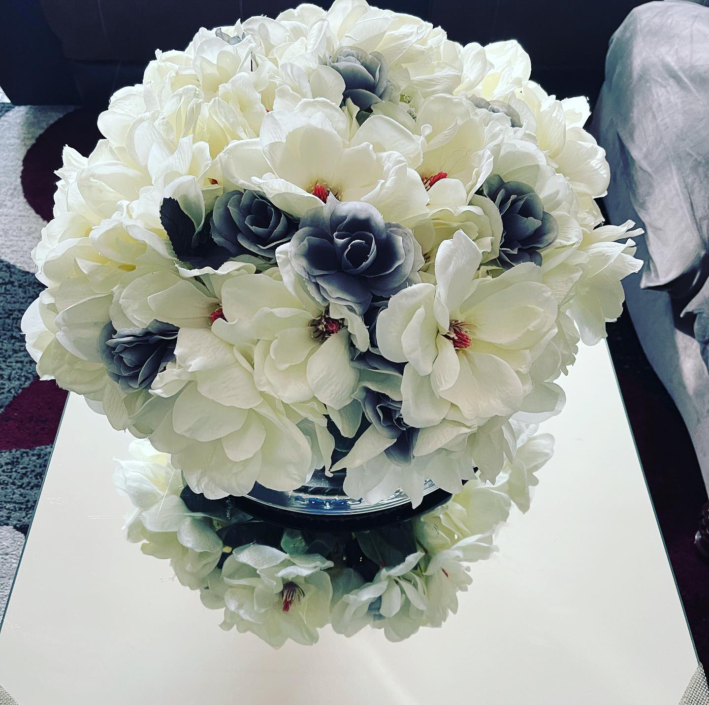 Round white and gray-blue silk rose centerpiece with green fern leaves on a mirrored tabletop reflecting the arrangement