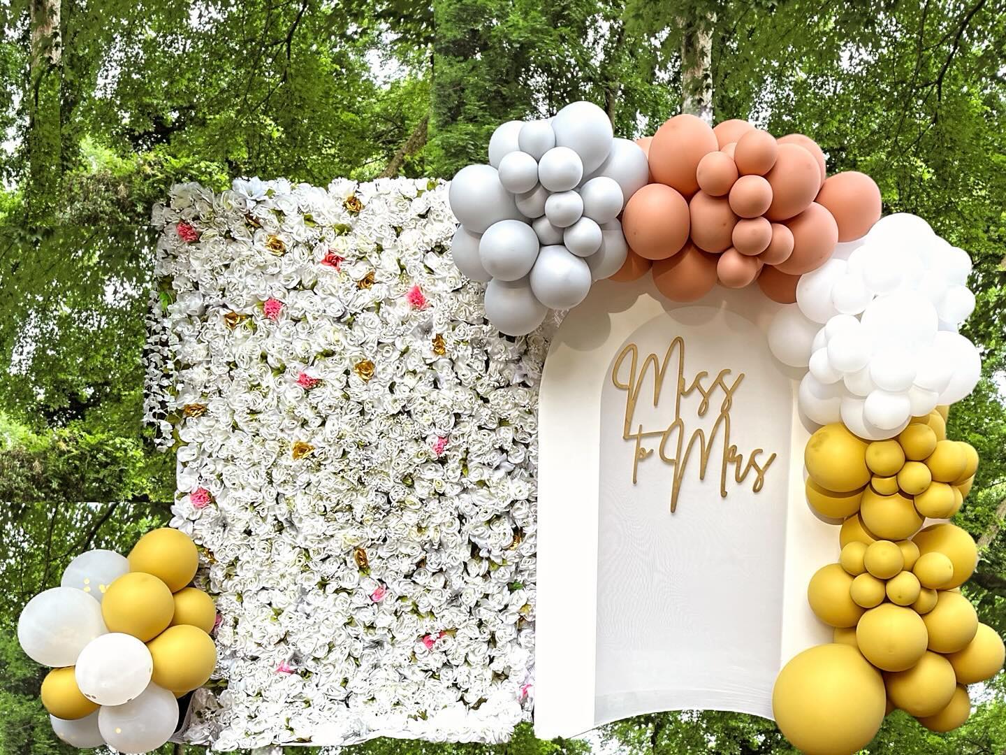 White flower wall backdrop paired with an arched panel reading a wedding-style monogram, framed by gray, tan, white and mustard balloon garlands