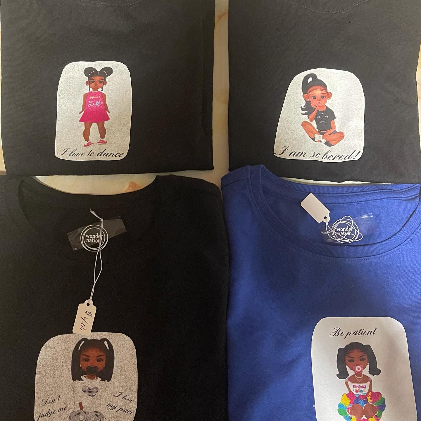 Flat lay of folded black and blue T-shirts with printed cartoon girl graphics and phrases like 'I love to dance'