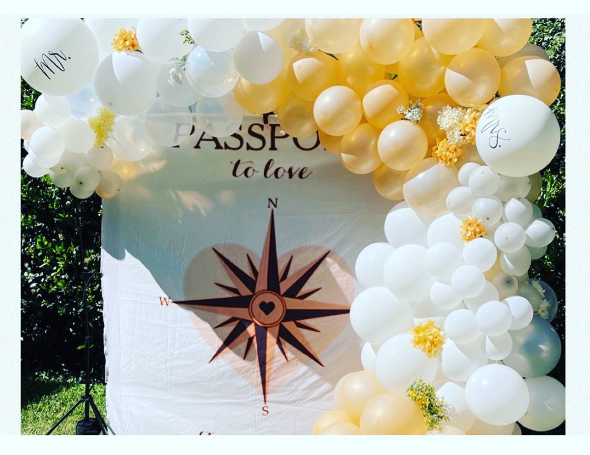 White backdrop reading Passport to Love with a compass and heart graphic, framed by white and gold balloon garland and small yellow flowers