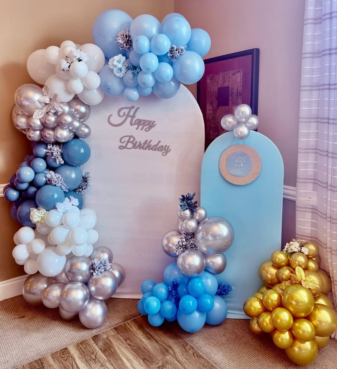 Balloon backdrop arch in blue, white and silver with floral accents and a Happy Birthday script sign, plus a separate gold balloon cluster