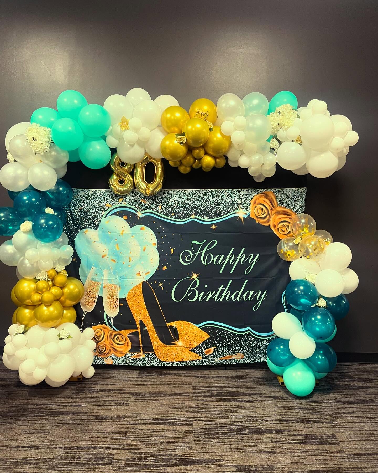 Black backdrop with gold '80' balloon numbers, teal, white and gold balloon garland, faux flowers, and a 'Happy Birthday' banner with an illustrated gold high heel and champagne glasses