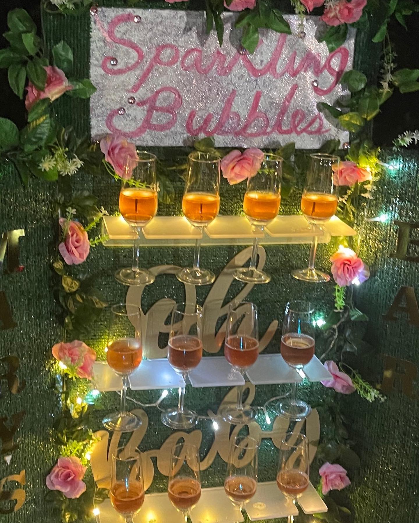 Green foliage backdrop with a 'Sparkling Bubbles' sign above tiered shelves holding filled champagne glasses and pink faux roses