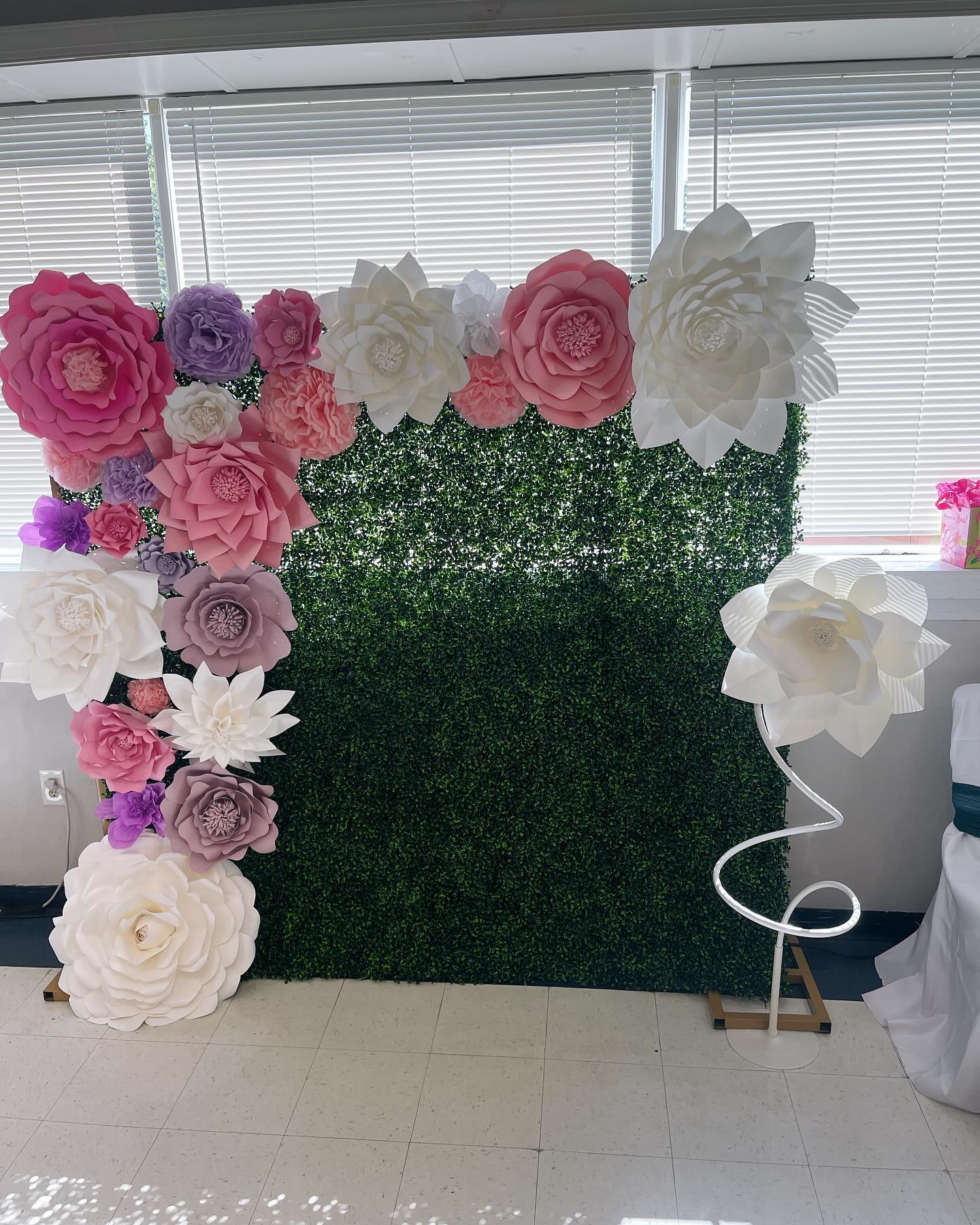 Large paper flower wall backdrop in pink, white and purple tones mounted on an artificial greenery panel with a standing paper flower light beside it
