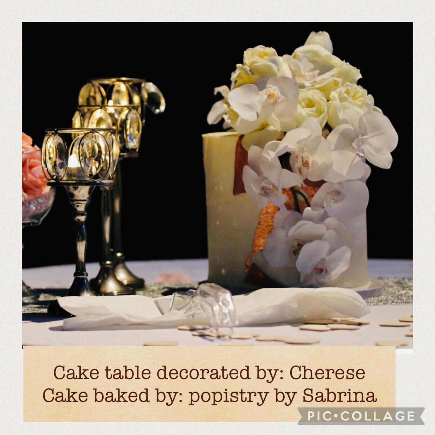 Close-up of a round cake decorated with cream roses and white orchids, beside ornate candle holders on a table with pink flowers.