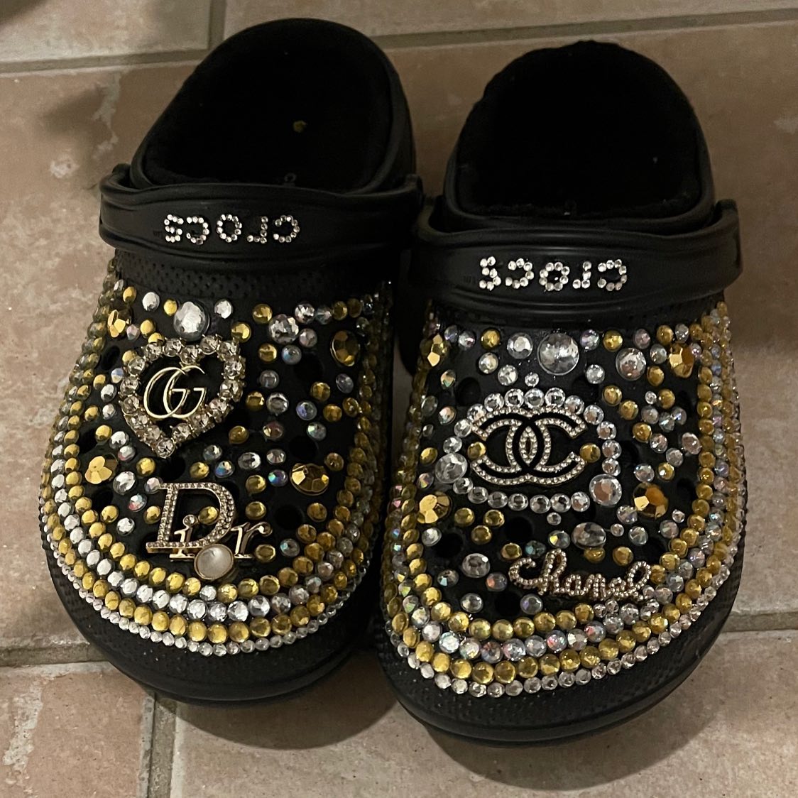 Pair of black clog sandals decorated with gold and silver rhinestones and designer-style charms on a tile floor