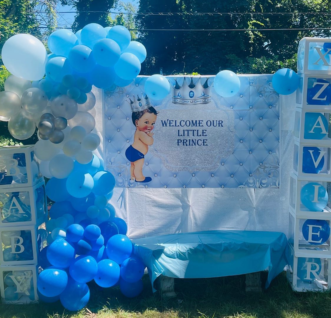 Blue and white baby shower backdrop reading Welcome Our Little Prince with an illustrated crowned baby, framed by blue and silver balloon garland and BABY block letters