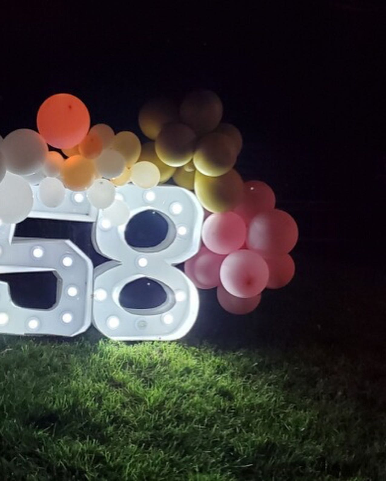 Light-up marquee numbers reading 58 decorated with orange, yellow, and pink balloon clusters, glowing at night on grass