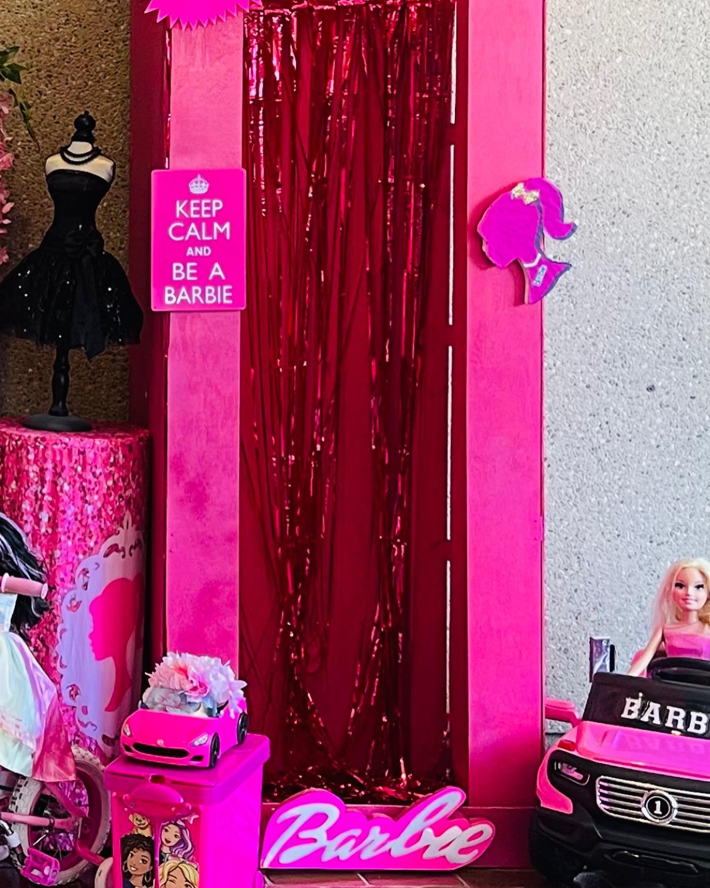 Close-up of a pink Barbie box photo prop with metallic fringe curtain, a doll on a mannequin, and toy cars and accessories nearby