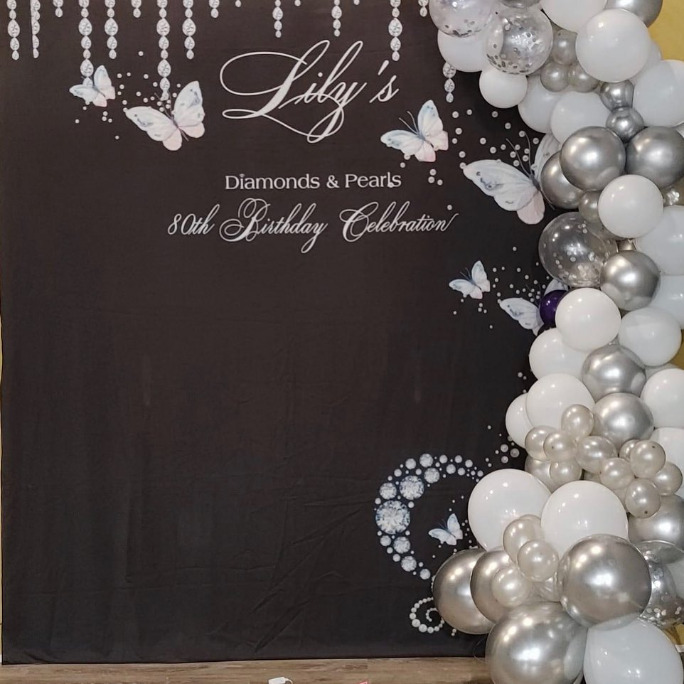 Black backdrop with silver butterfly and diamond graphics reading 'Diamonds & Pearls 80th Birthday Celebration' beside a white and silver balloon garland