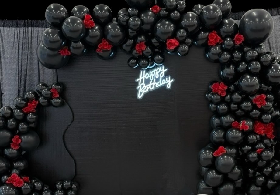 Black balloon garland backdrop with red rose accents and a glowing Happy Birthday neon sign against a dark curtain