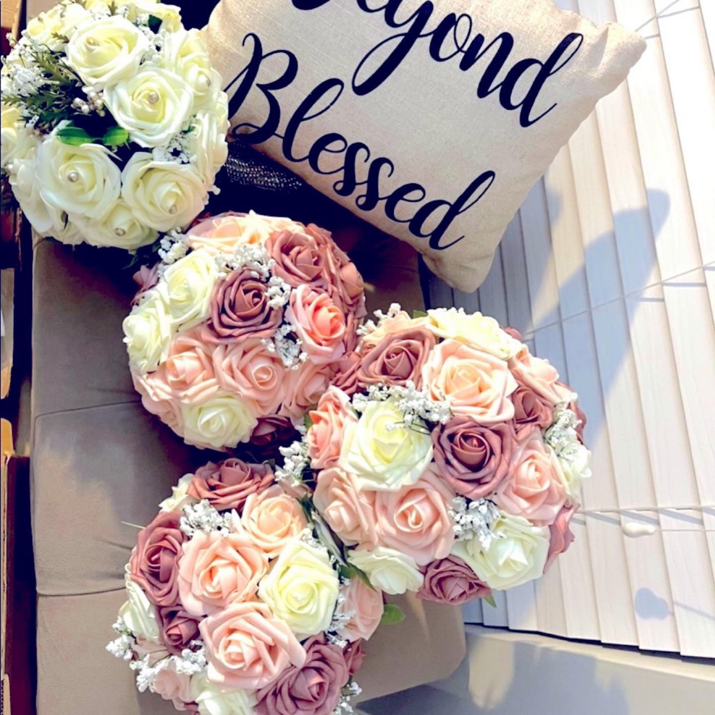 Cream and dusty-pink faux rose bouquets arranged on a tan tufted bench beside a pillow reading 'Beyond Blessed'