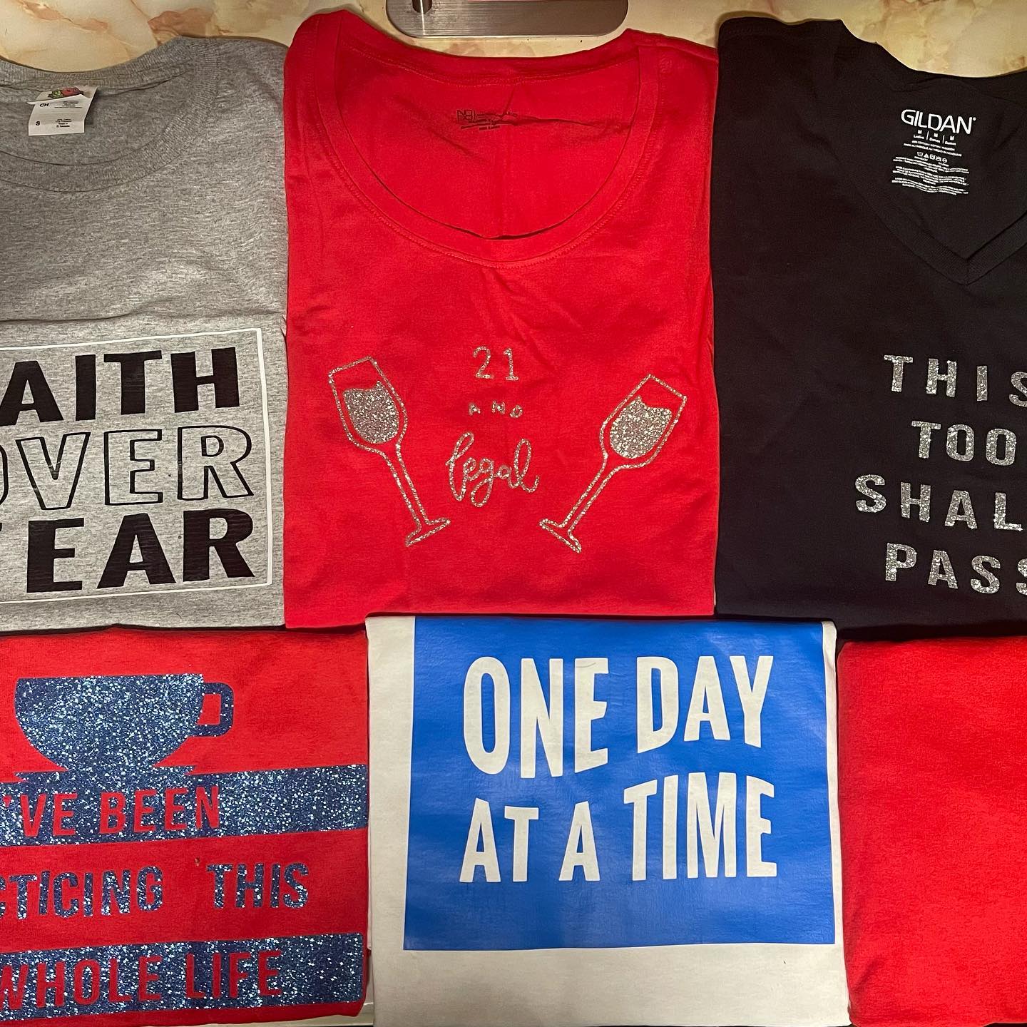 Flat lay of folded T-shirts including grey, red, black, and blue designs with slogans like 'One Day At A Time'