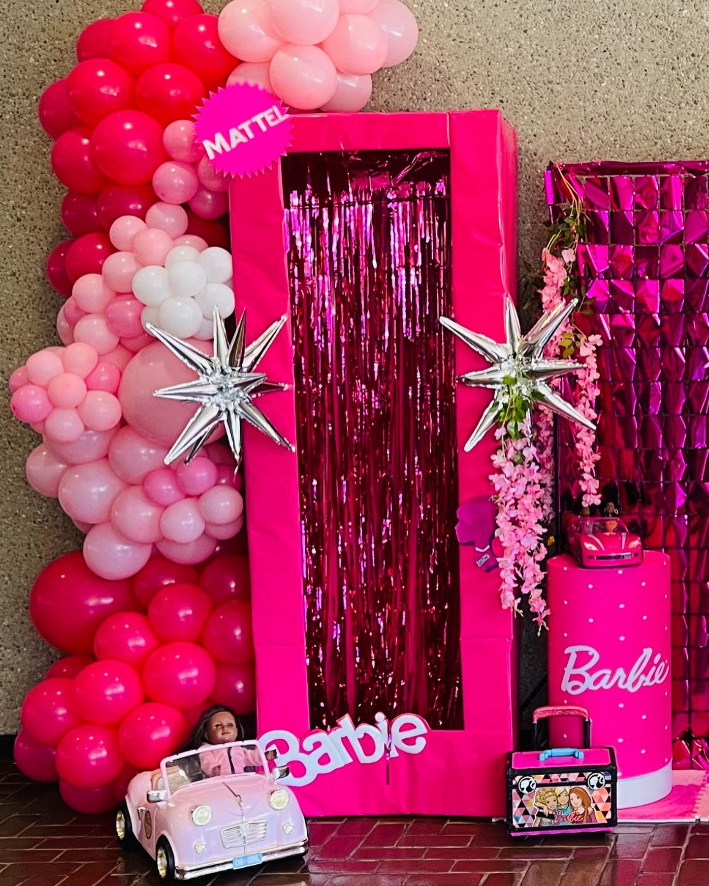 Life-size pink Barbie box display with metallic fringe curtain, silver star balloons, floral garland, and a toy car and lunchbox at the base