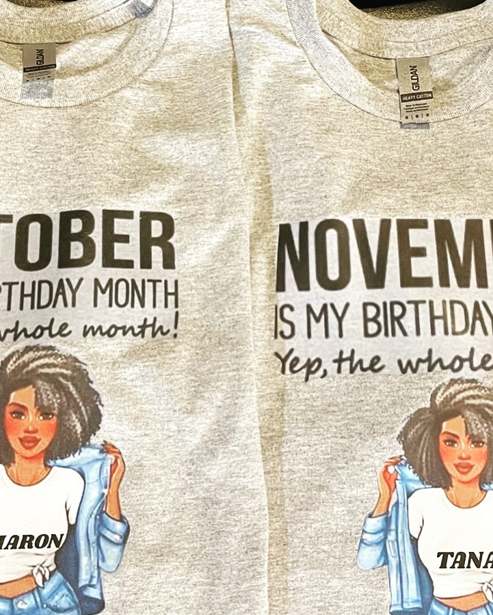 Two heather grey T-shirts on hangers printed with illustrated women and birthday-month text