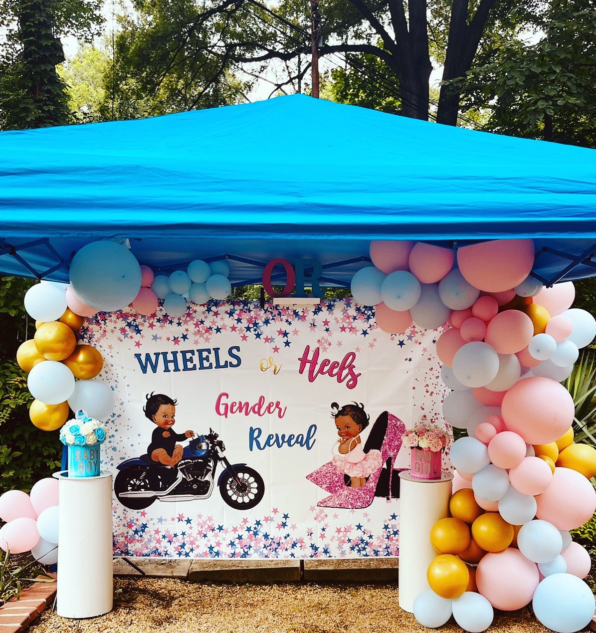White backdrop reading Wheels or Heels Gender Reveal under a blue pop-up tent, with illustrated cartoon babies on a motorcycle and in heels, flanked by balloon garland