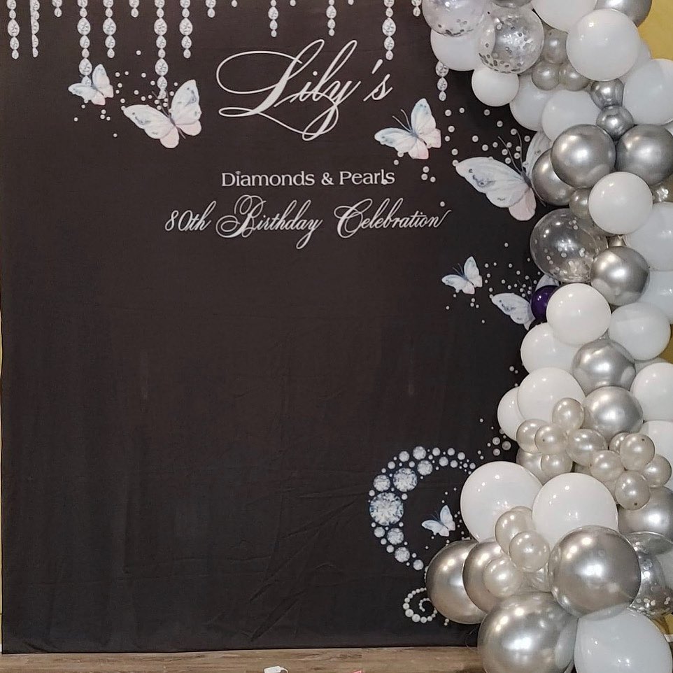 Black backdrop with silver butterfly and diamond graphics reading 'Diamonds & Pearls 80th Birthday Celebration' beside a white and silver balloon garland
