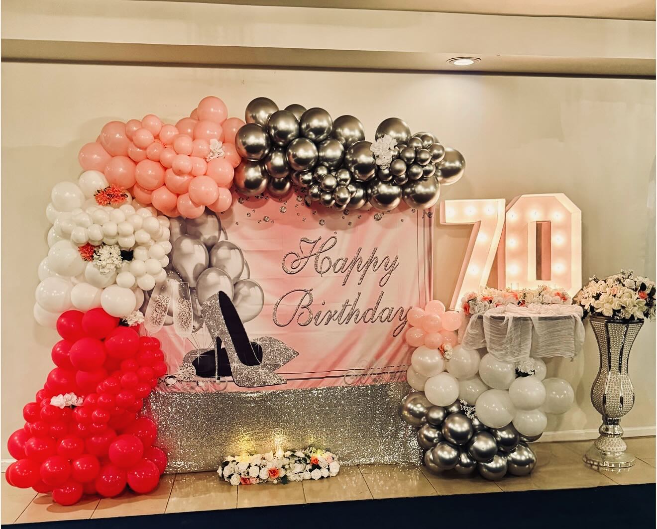 Backdrop display for a 70th birthday featuring a Happy Birthday banner, glitter high heel graphic, marquee light-up 70 numbers, and coral, silver and red balloon garlands