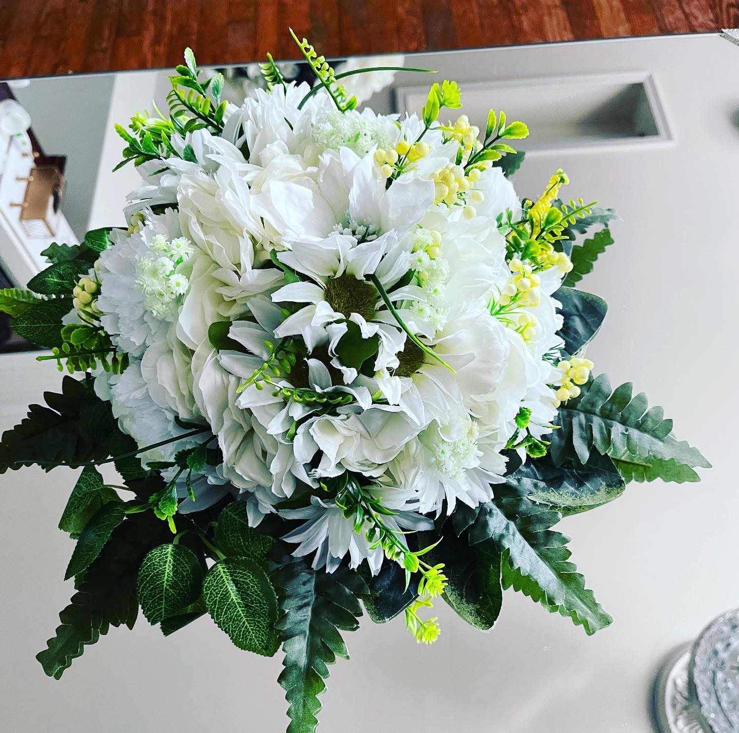 Round white and yellow silk flower centerpiece with ferns and greenery on a mirrored tabletop