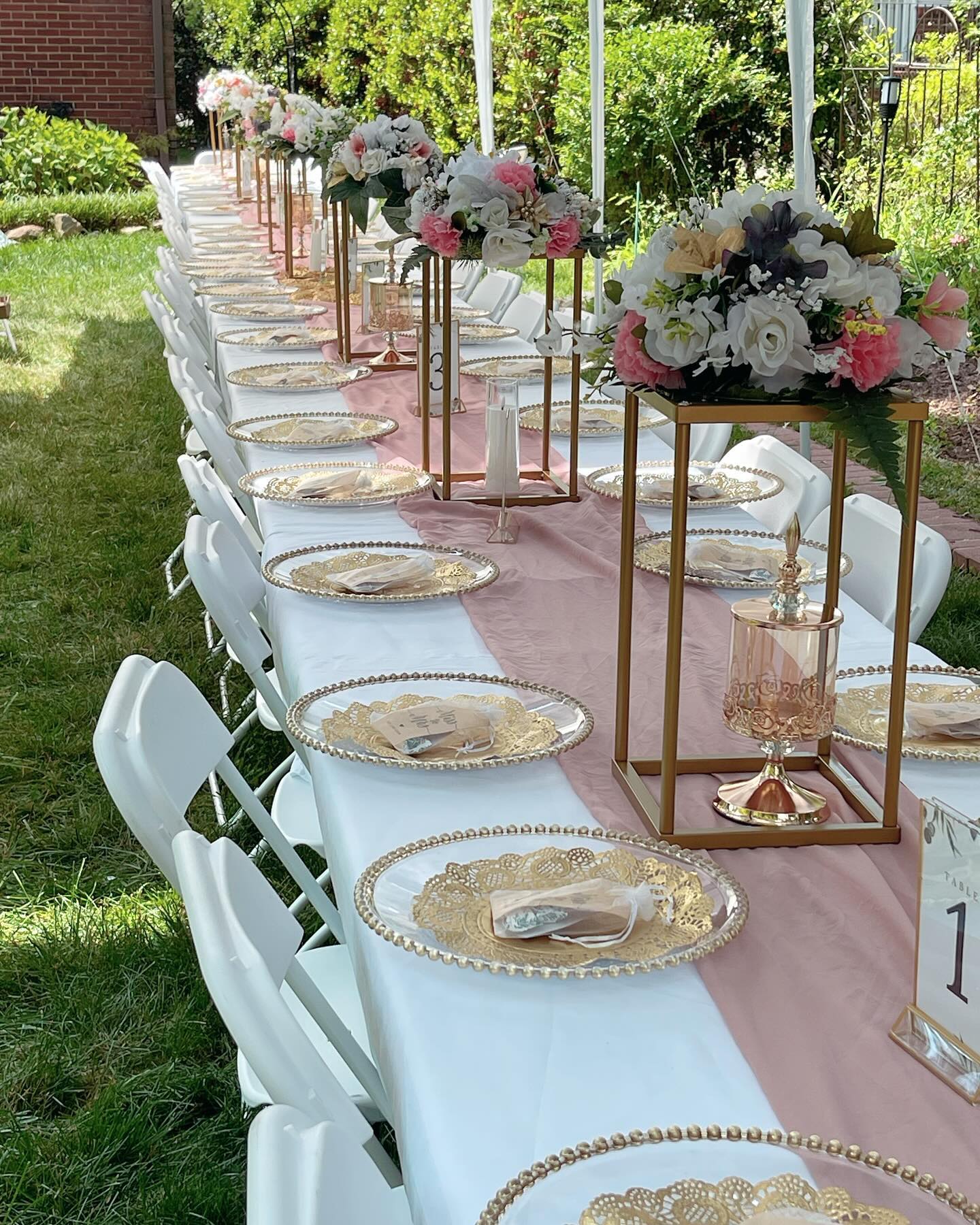 Long outdoor banquet table with gold beaded chargers, tall floral centerpieces on gold stands, and a pink table runner under a tent