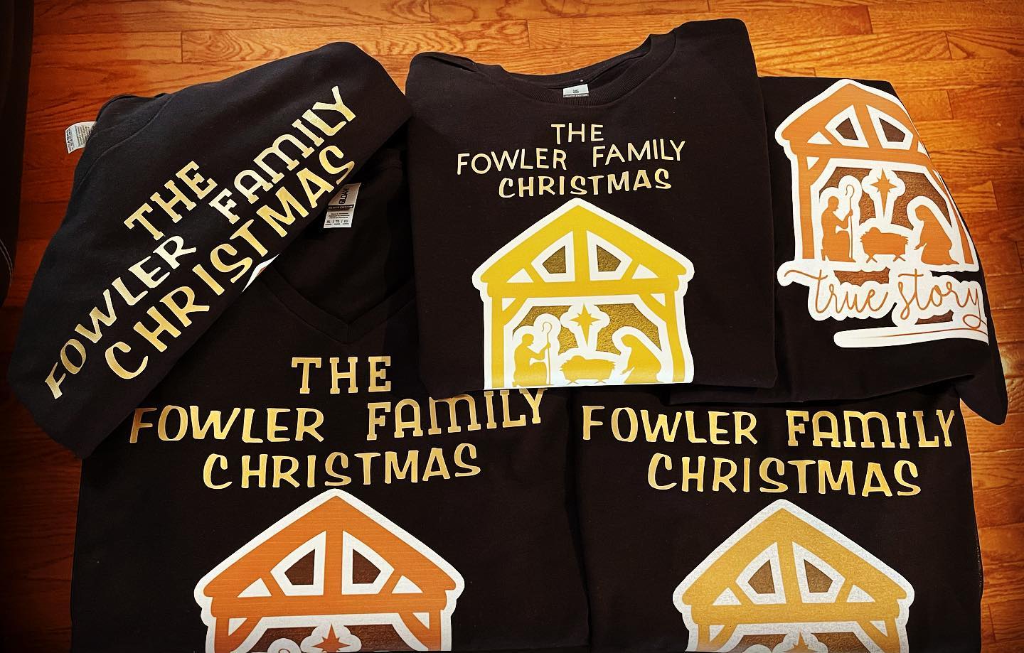 Stack of folded black t-shirts and sweatshirts printed with gold and orange nativity stable graphics and holiday text on a wood floor