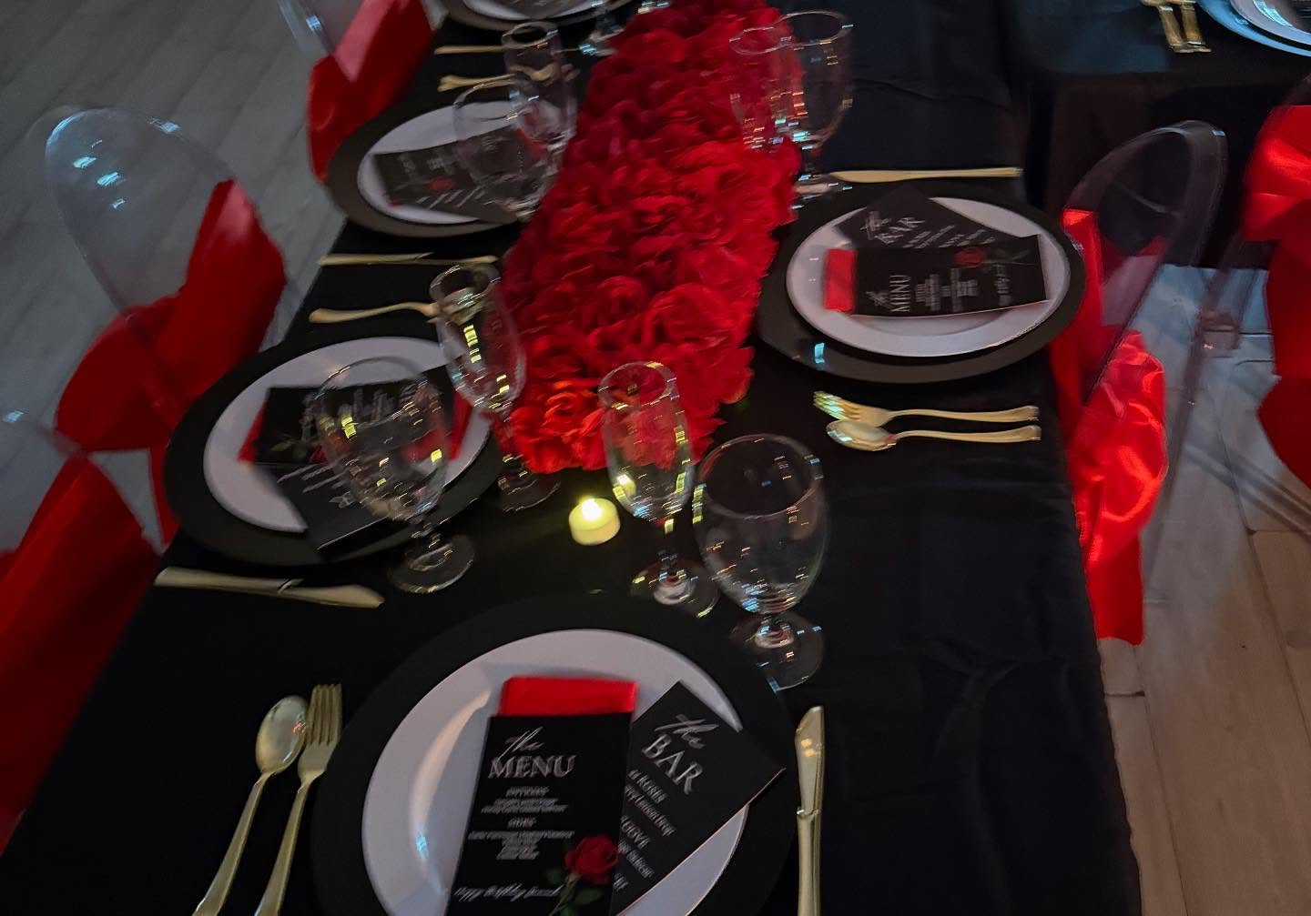 Formal table setting with black chargers, menu and bar cards, a red rose runner, gold flatware, candle, and red satin chair bows