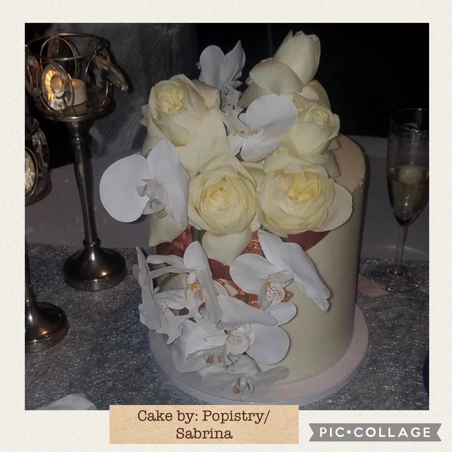 Close-up of a round white cake topped with cream roses and white orchid decorations, beside a champagne glass and candle holders.