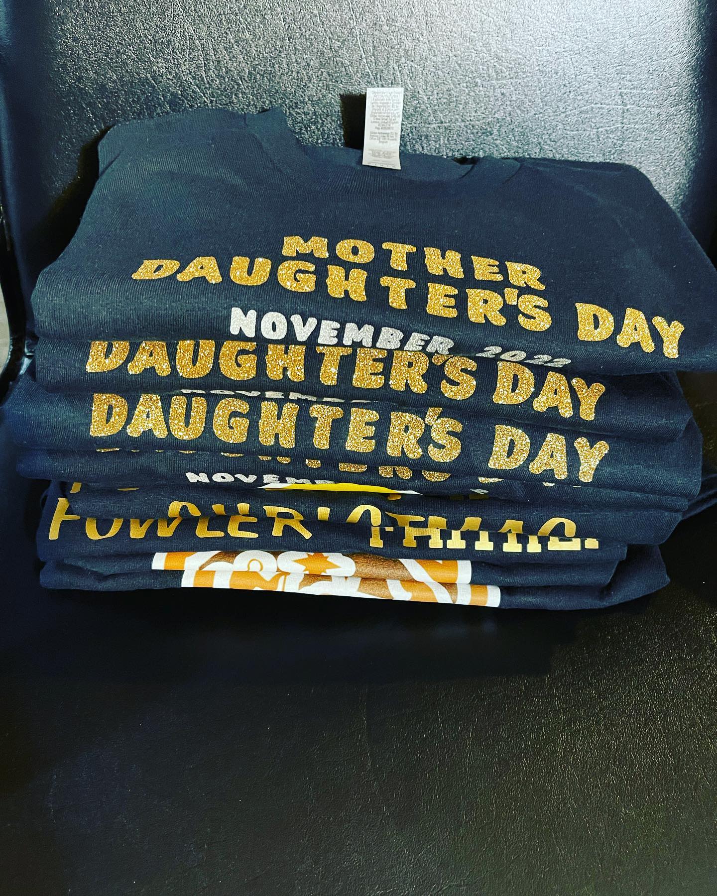 Stack of folded navy t-shirts printed with gold glitter text about a mother-daughter event and date, with an orange and white graphic shirt underneath