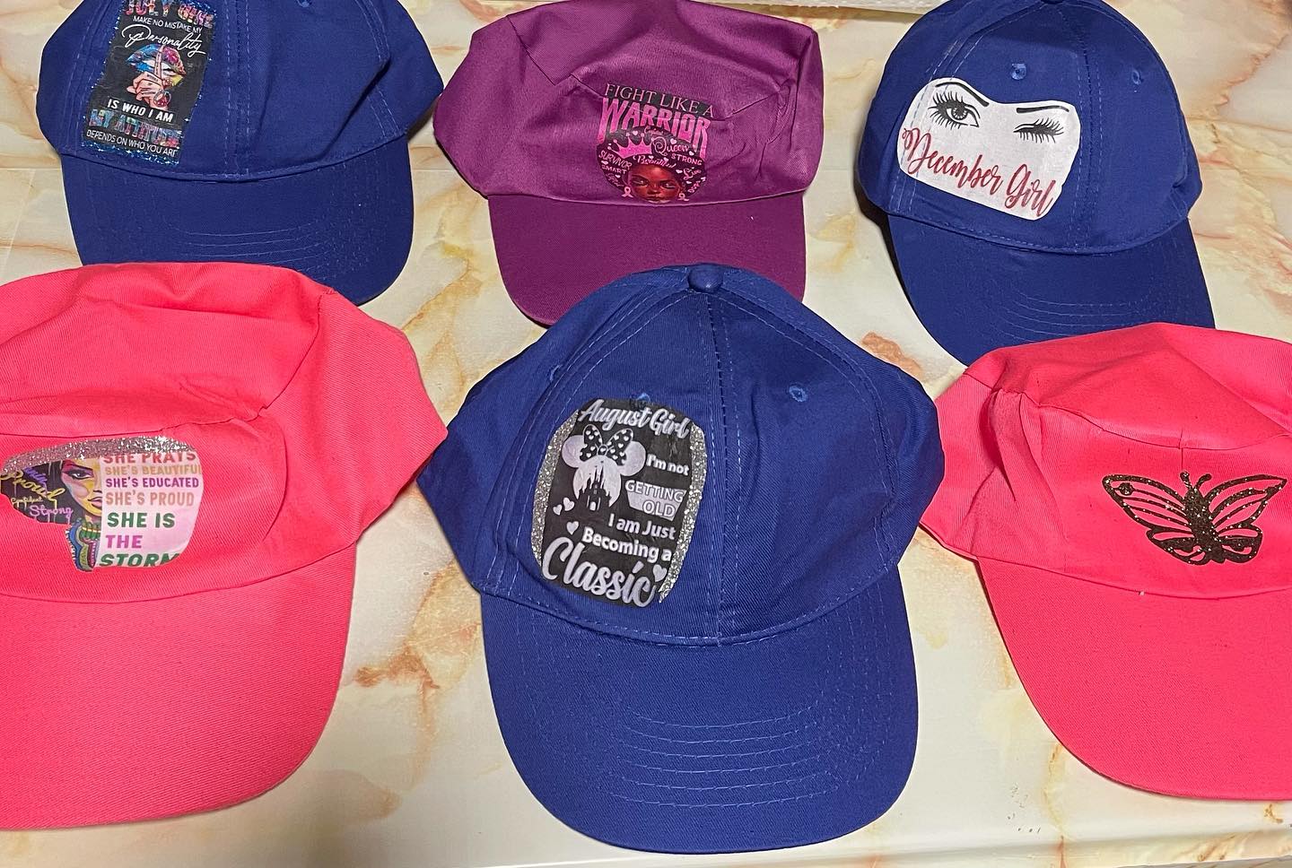 Six custom baseball caps in blue and pink with printed graphics and slogans like 'August Girl' and 'December Girl'