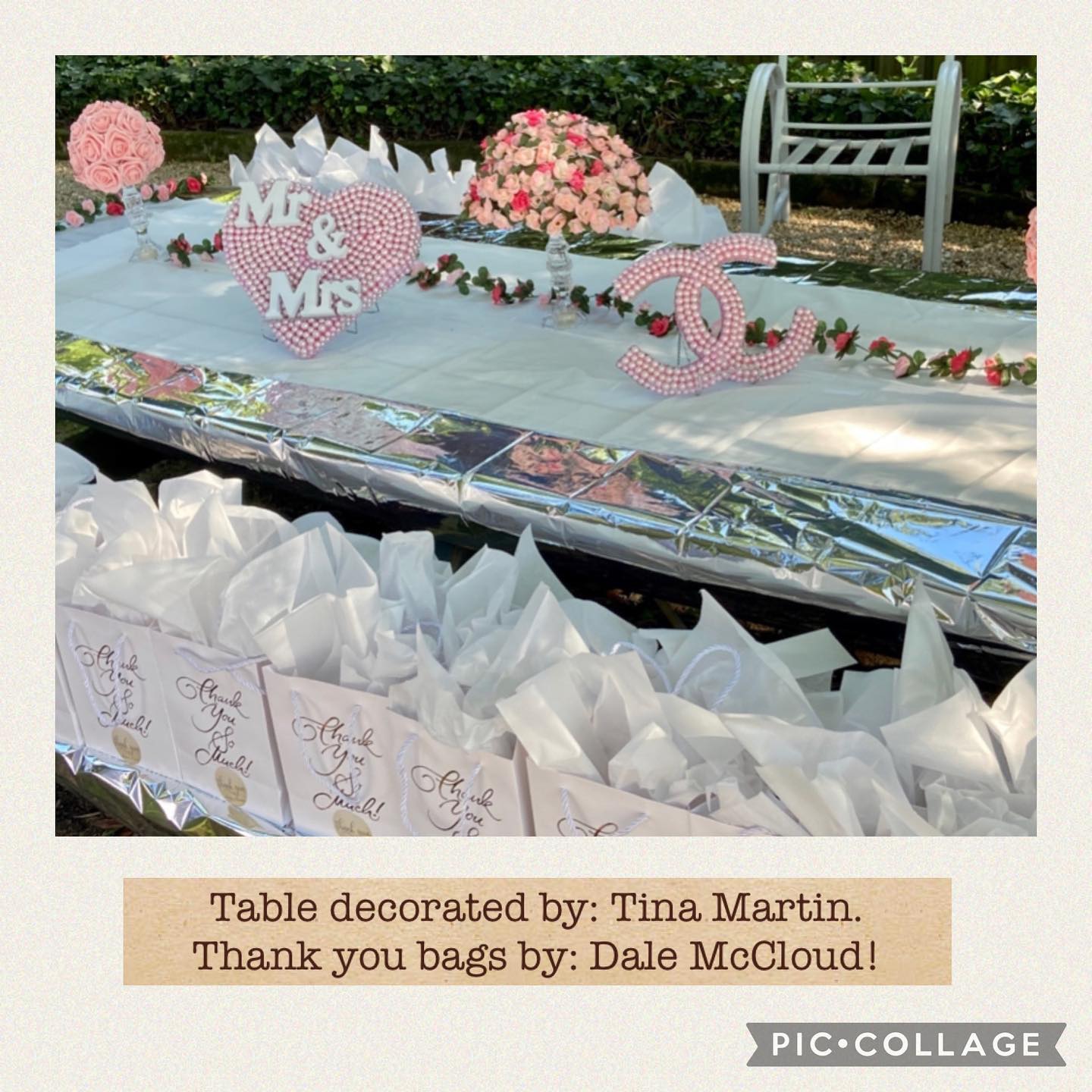 A long table displays a beaded heart 'Mr & Mrs' sign, an interlocking-ring monogram sign, pink floral arrangements, and white gift bags with tissue paper.